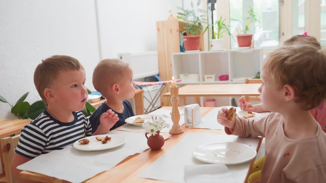 Bambino con apparecchio acustico che si protende verso la candela durante il dessert, spingendo un vaso di fiori mentre cerca di spegnere la luce; altri siedono mangiando un dolce a un tavolo in una luminosa aula di scuola materna, un momento di curiosità