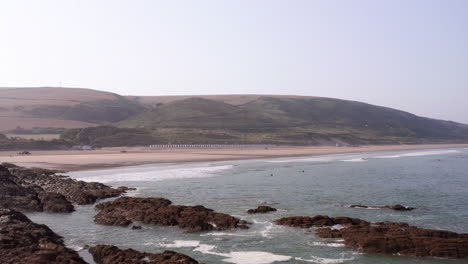 Sobrevuelo-Aéreo-De-Olas-Rompiendo-En-Rocas-Con-Una-Playa-De-Arena-En-El-Fondo