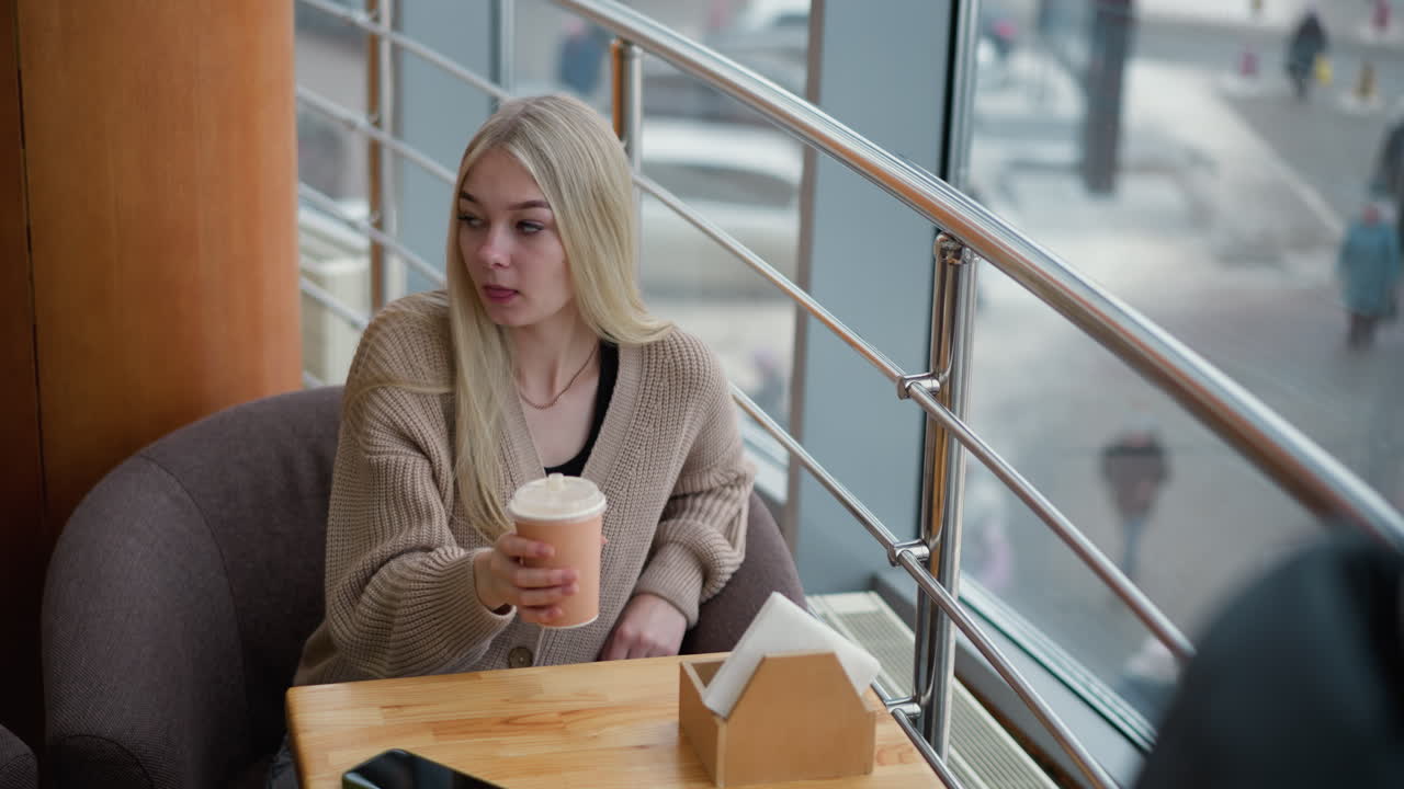 Jovem mulher tomando chá de sua xícara em um café aconchegante, acidentalmente derrubando-a sobre a mesa de madeira, enquanto o fundo mostra pessoas se movimentando em uma cena de inverno agitada do lado de fora da janela do café.
