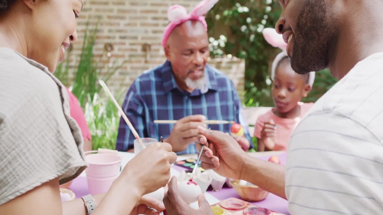 幸せそうなアフリカ系アメリカ人の両親、娘、祖父が庭でイースターエッグに色を塗っている、スローモーション