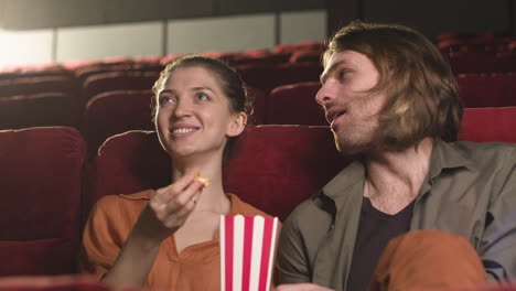 happy couple sitting in the cinema while they talking and eating popcorn waiting fot watch the film 1