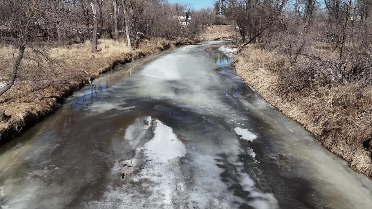Kışın güney Manitoba'daki Rat Nehri üzerinde alçaktan uçan drone ile kenarlara yakın açık suyla eriyen buzları, yerdeki karı ve her iki yandaki ormanı gösteren görüntü