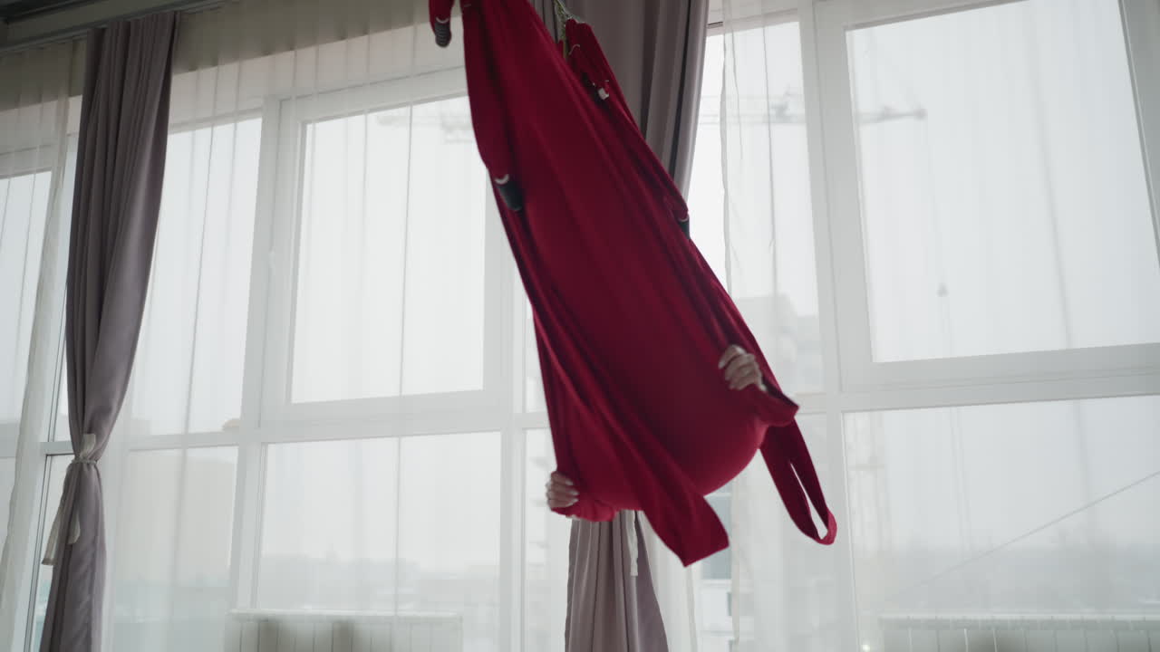 Bright yoga studio with floor to ceiling windows and sheer curtains as spiritual mentor lies comfortably in red silk hammock with eyes closed smiling, swinging gently to convey calm wellness practice