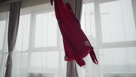 bright yoga studio with floor to ceiling windows and sheer curtains as spiritual mentor lies comfortably in red silk hammock with eyes closed smiling, swinging gently to convey calm wellness practice