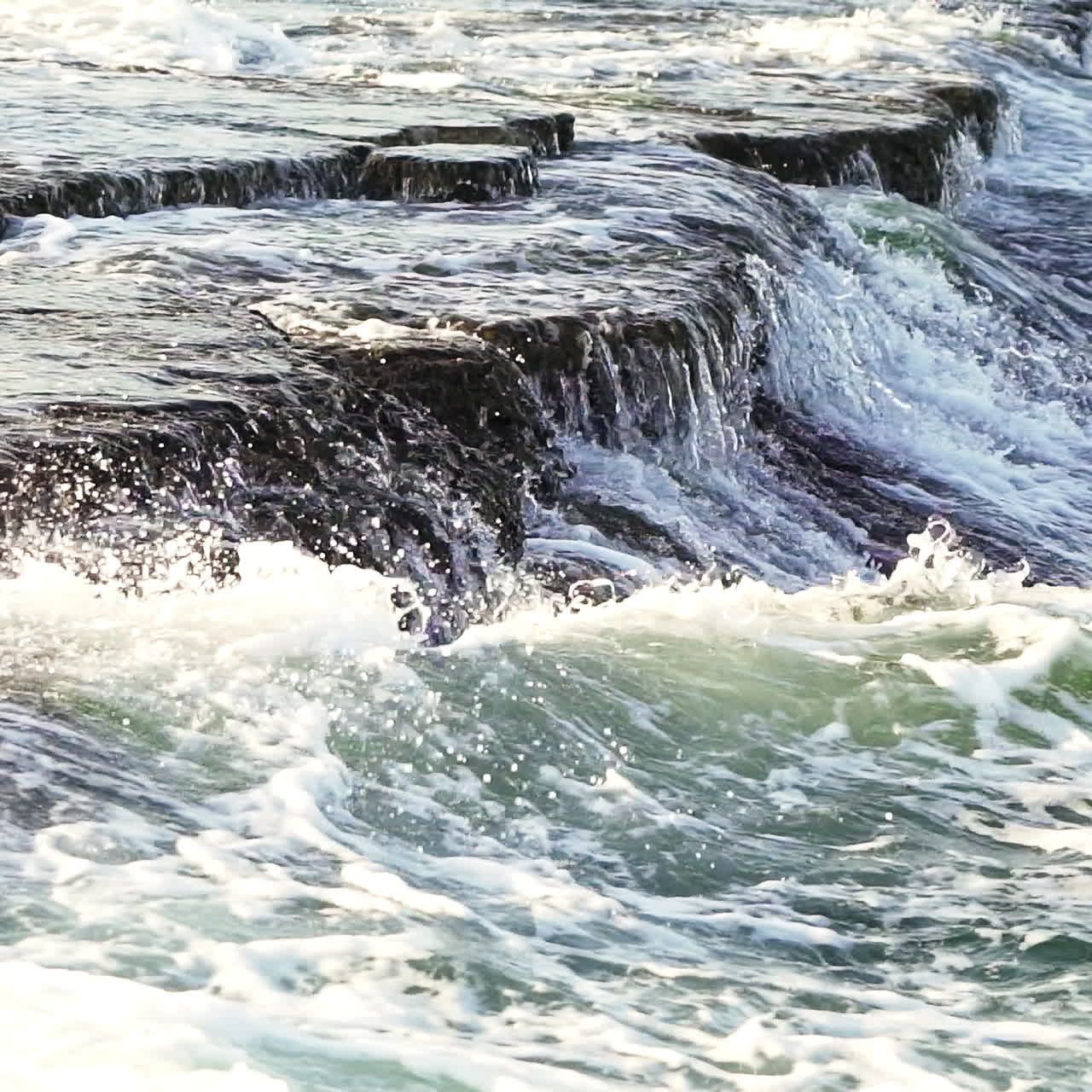 Water flowing over the rocky surface and waves are making white foam. Powerful ocean wave splashes across many rocks. Breathtaking view of breaking wave foaming on black rocks.