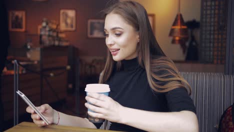 hermosa joven sentada a la mesa en el café con una taza de café en las manos y navegando por el teléfono inteligente