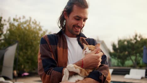 portrait: a brunette guy with long hair holds a red cat and strokes it. rest in the country house