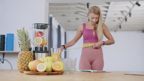 blonde girl uses measuring tape on body next to fruit and mixer, slomo