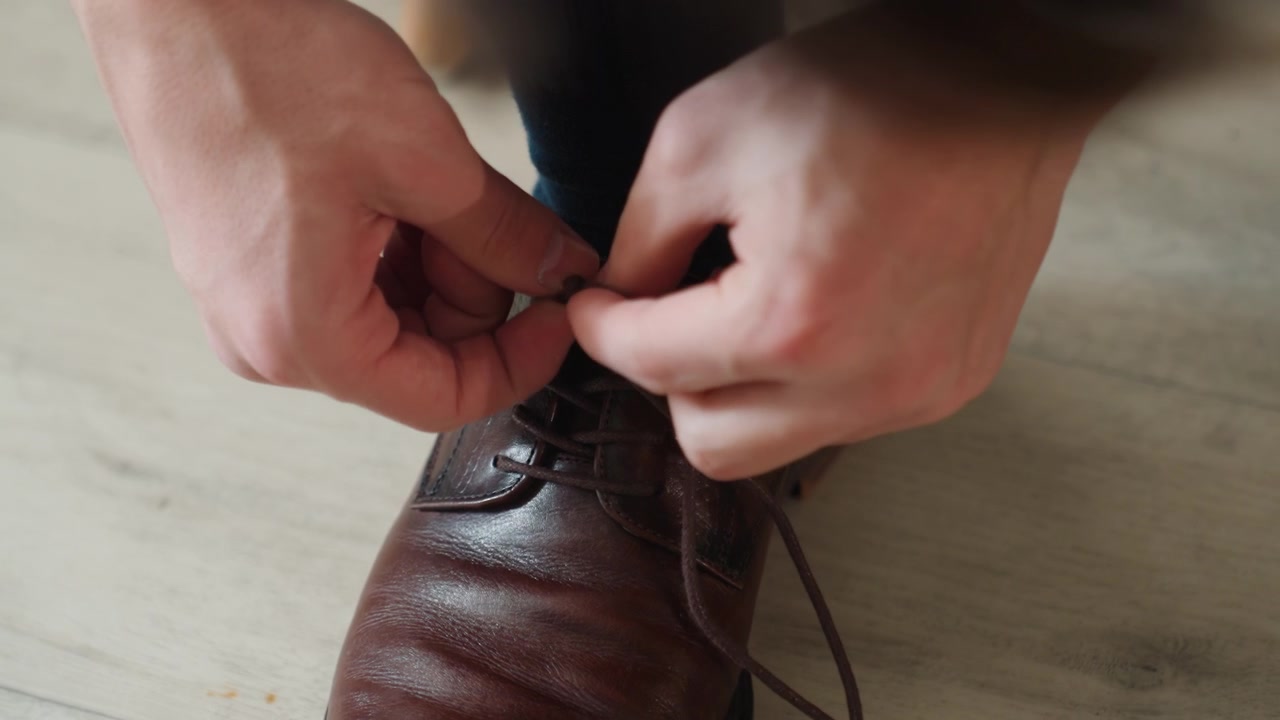 Primo piano ravvicinato di una scarpa oxford marrone che viene allacciata, mani svelte che stringono i lacci sulla pelle lucida prima della routine per il pendolarismo, uno stile pronto per l'ufficio, nodi veloci e una presa salda sul pavimento in legno, trasmette