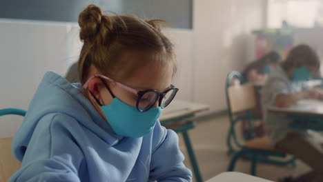 Girl-in-face-mask-learning-in-classroom.-Schoolgirl-writing-in-notebook