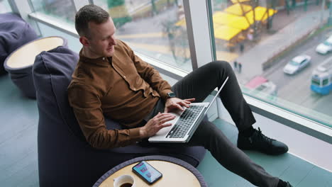 Young freelancer uses laptop. Handsome guy is sitting by the window with city view background and working on business with a wireless computer.