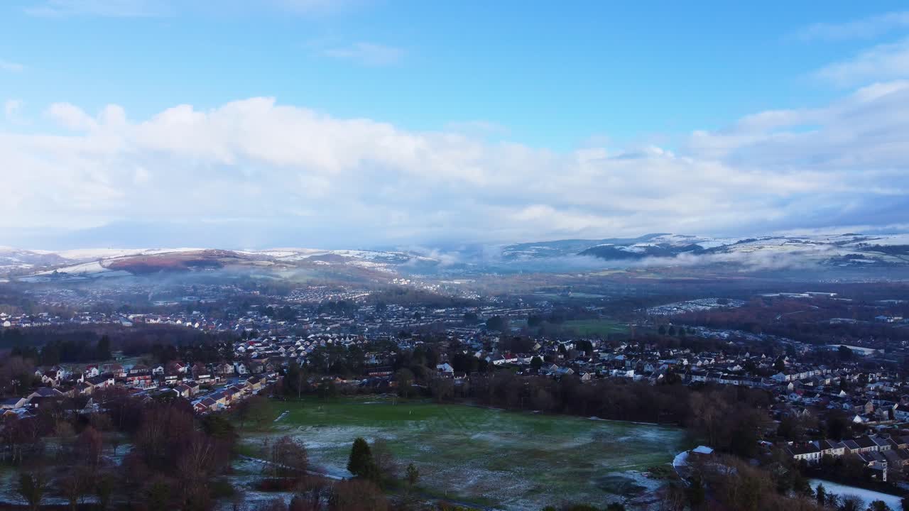 Luftaufnahmen über einem Park mit umliegenden Häusern und Gebäuden, wobei Schnee das höher gelegene Gelände bedeckt. Perfekt für Naturdokumentationen und Winterreise-Promos. Gefilmt im Winter in Swansea, Großbritannien.