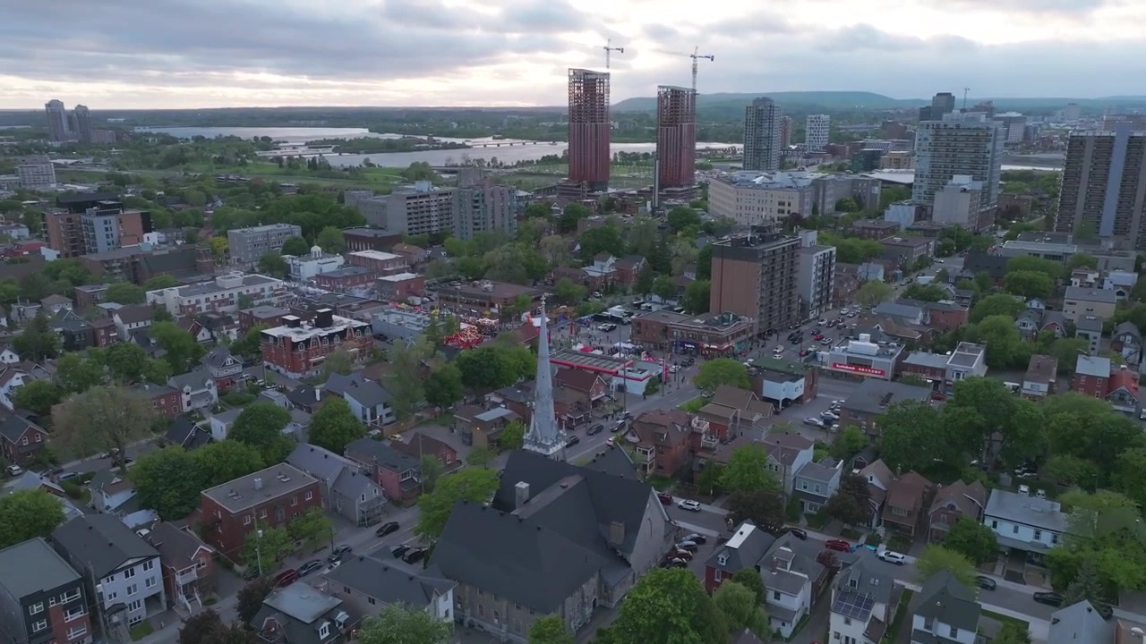 Una toma de dron de gran angular y picado que muestra un barrio residencial urbano de Ottawa, destacando una prominente aguja de iglesia, densos árboles verdes, rascacielos y el río Ottawa bajo un cielo nublado.