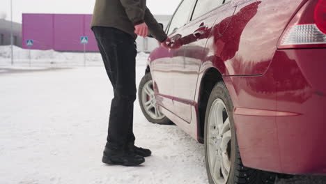 factory worker in winter clothing walks across snowy ground toward red car in open parking lot with snow-covered surface, distant industrial buildings, road signs, and leafless trees in background
