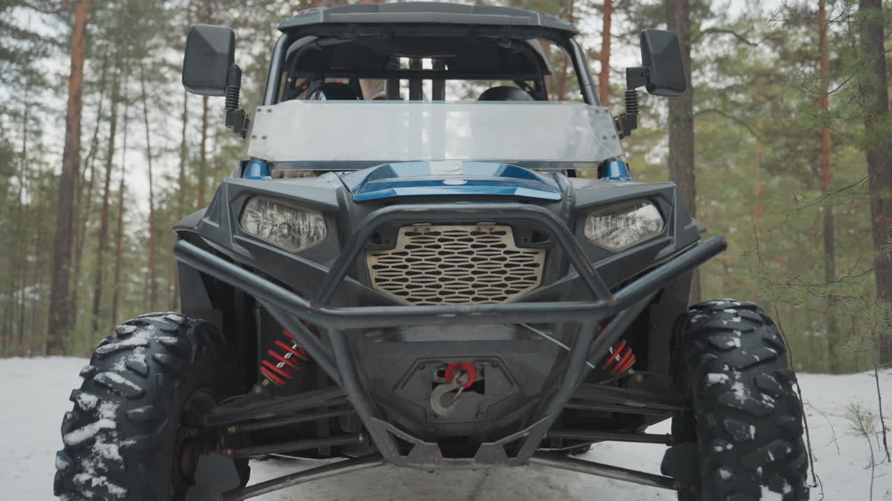 Quad in una pineta innevata, veicolo utilitario robusto incorniciato da alte conifere, paraurti rinforzato con verricello visibile, pneumatici con battistrada aggressivo ricoperti di neve, griglia anteriore e fari illuminati.