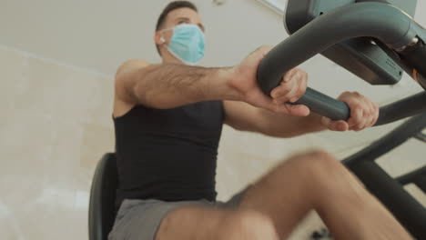 joven atleta fuerte hombre con mascarilla usa una máquina de ejercicio y bebe agua en el gimnasio