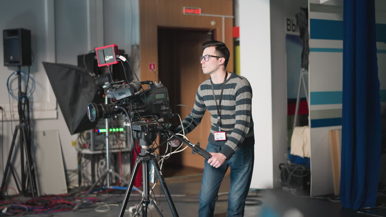 Man operating professional camera, adjusting camera controls, focusing on video production in studio with equipment and colorful background, preparing for filming with clear concentration