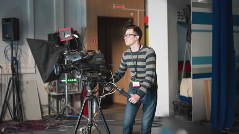 man operating professional camera, adjusting camera controls, focusing on video production in studio with equipment and colorful background, preparing for filming with clear concentration