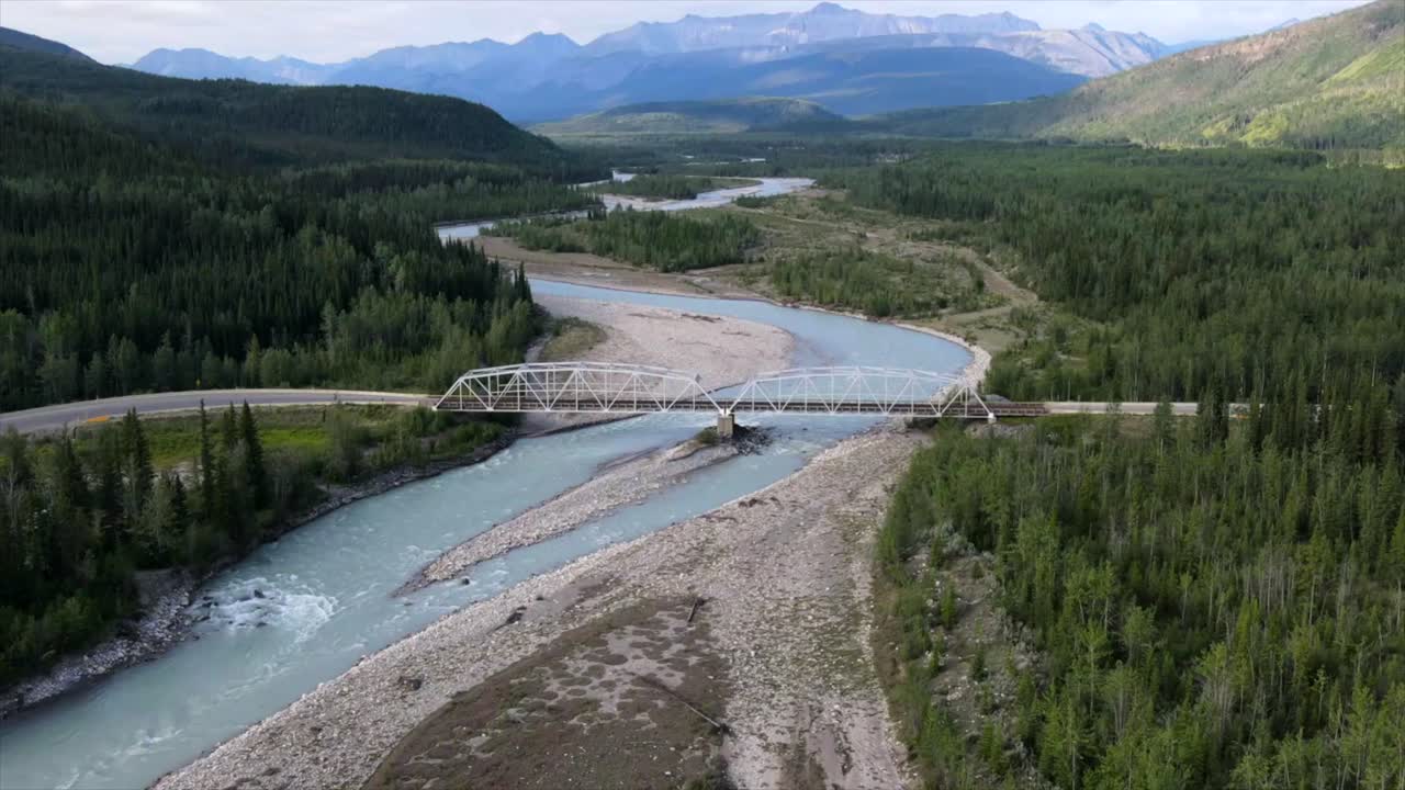 Aerial Hyperlapse of glacier river running through valley between beautiful mountains and lush endless forests. Turquoise water flowing underneath old steel bridge with cars driving across.