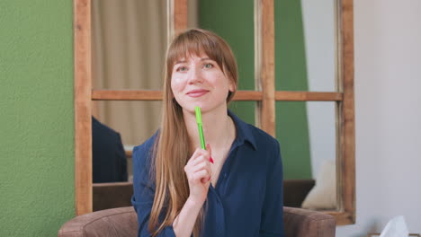 therapist seated on sofa writing with green pen on notepad, lifting face with calm smile in cozy room featuring green wall, wooden framed mirror behind her, pen holder and tissue box on nearby table