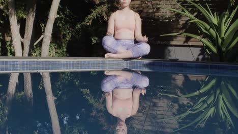 Focused-biracial-woman-practicing-yoga-and-meditating-at-pool-in-garden-in-slow-motion