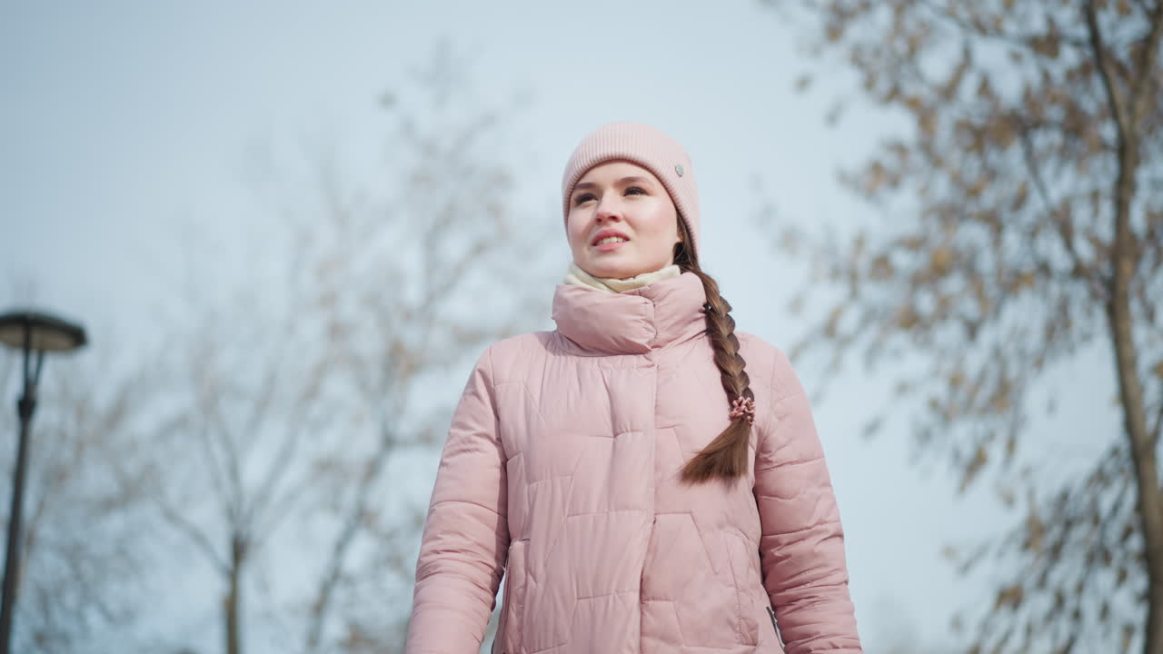 Young woman in pink winter jacket and hat standing outdoors on cold day, looking up into distance with soft expression, surrounded by leafless trees and clear sky, evoking seasonal change and peaceful reflection