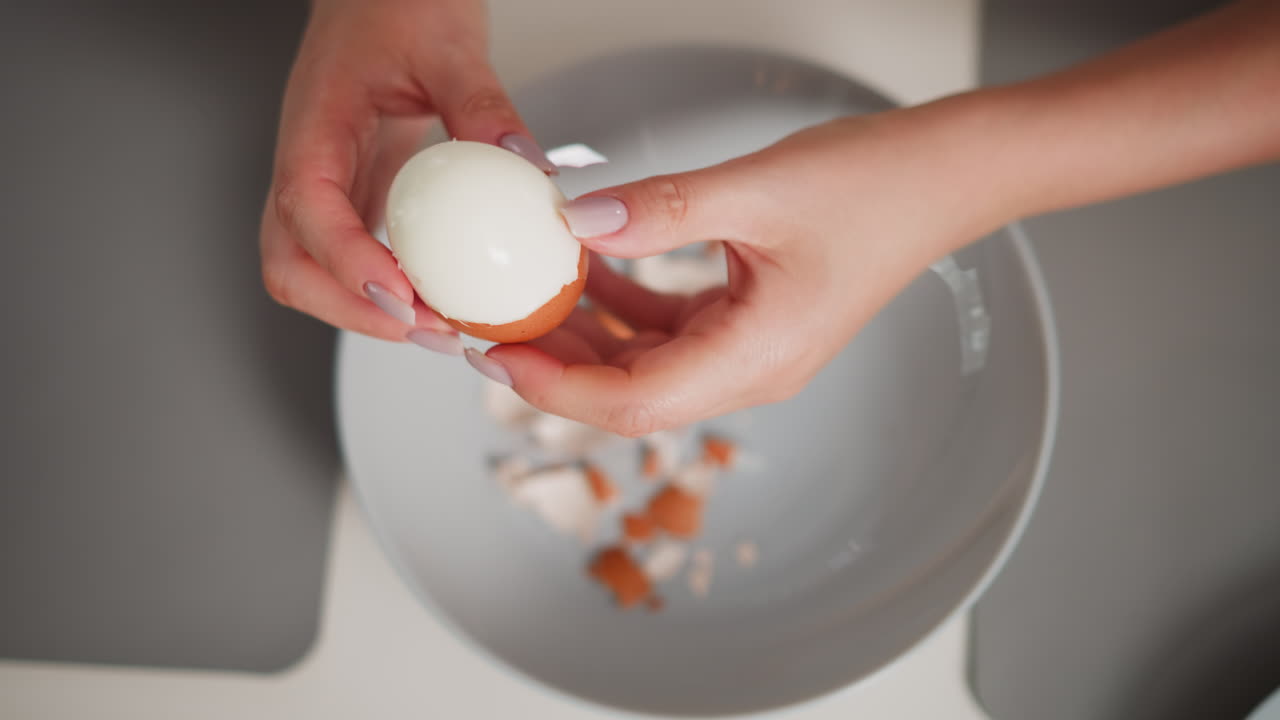 Close up aerial view of fair skinned person peeling shell from boiled egg over bowl filled with broken egg shell pieces, both hands carefully working in bright kitchen setting with grey placemats