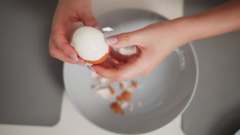 close up aerial view of fair skinned person peeling shell from boiled egg over bowl filled with broken egg shell pieces, both hands carefully working in bright kitchen setting with grey placemats