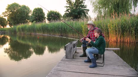 Side-view-of-a-teen-boy-sitting-with-his-grandfather-on-the-lake-pier,-talking-and-fishing-together