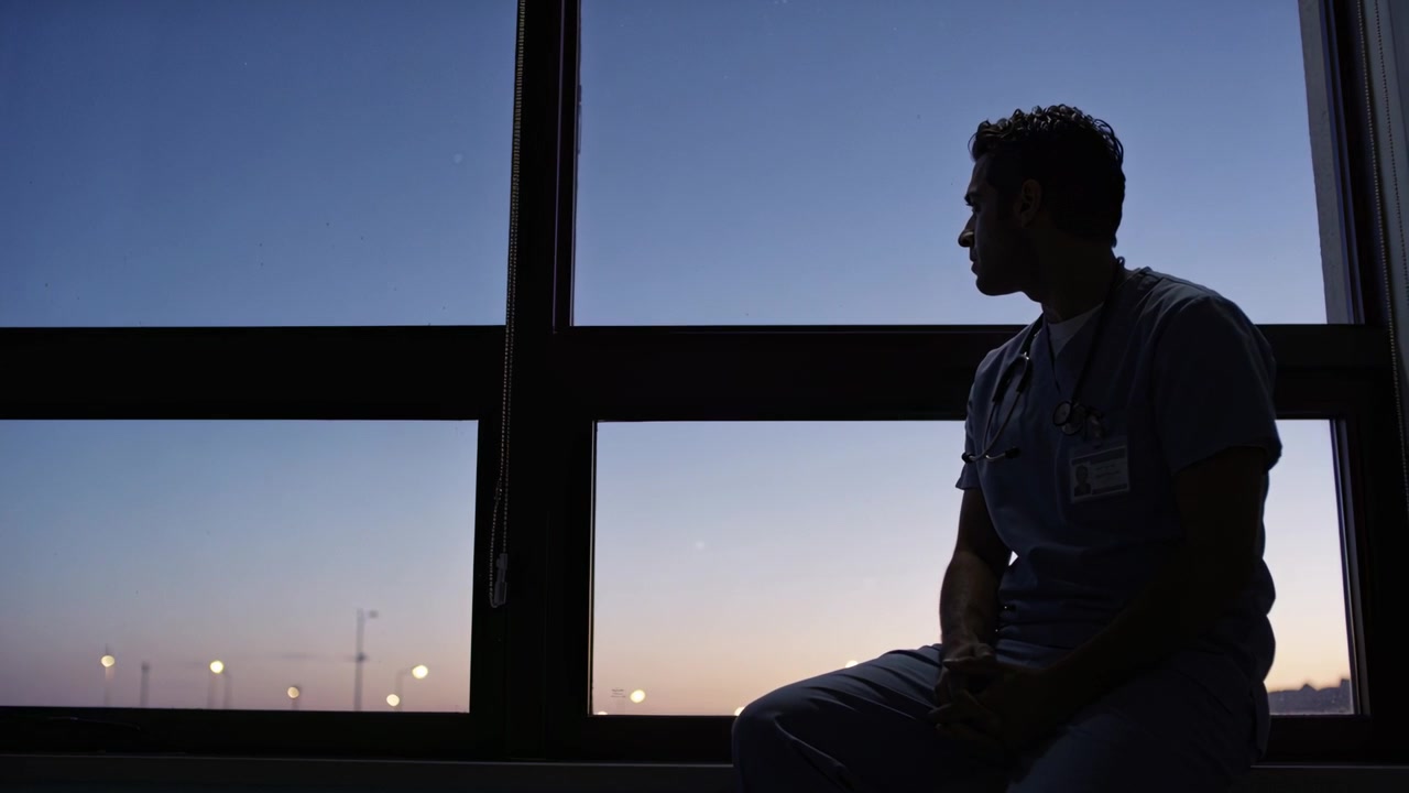 Shifting male doctor turning, gazing from sill as dusk settles, in scrubs, stethoscope, copy space