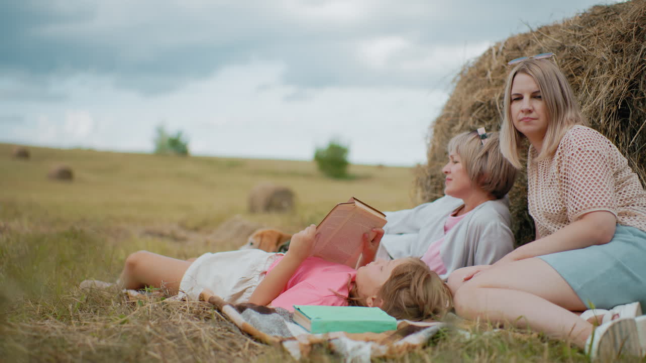 Family spending quality time together outdoors, relaxing on blanket in vast farmland, mother and daughter read books, dog resting beside them