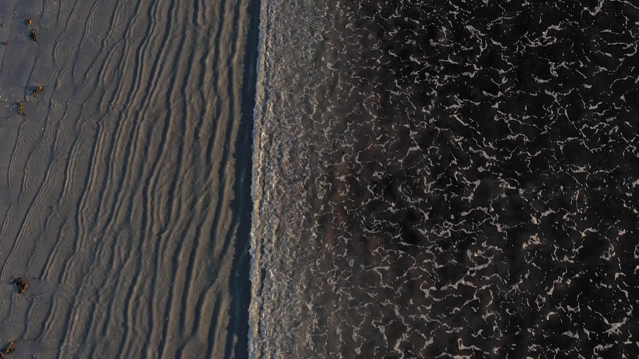 Aerial TOP DOWN flying over a series of waves coming in on a grey sand beach with cool patterns in the sand and surf