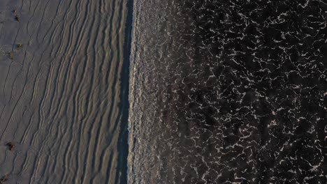 Antenne-Von-Oben-Nach-Unten-Fliegend-über-Eine-Reihe-Von-Wellen,-Die-An-Einem-Grauen-Sandstrand-Mit-Coolen-Mustern-Im-Sand-Und-In-Der-Brandung-Hereinkommen