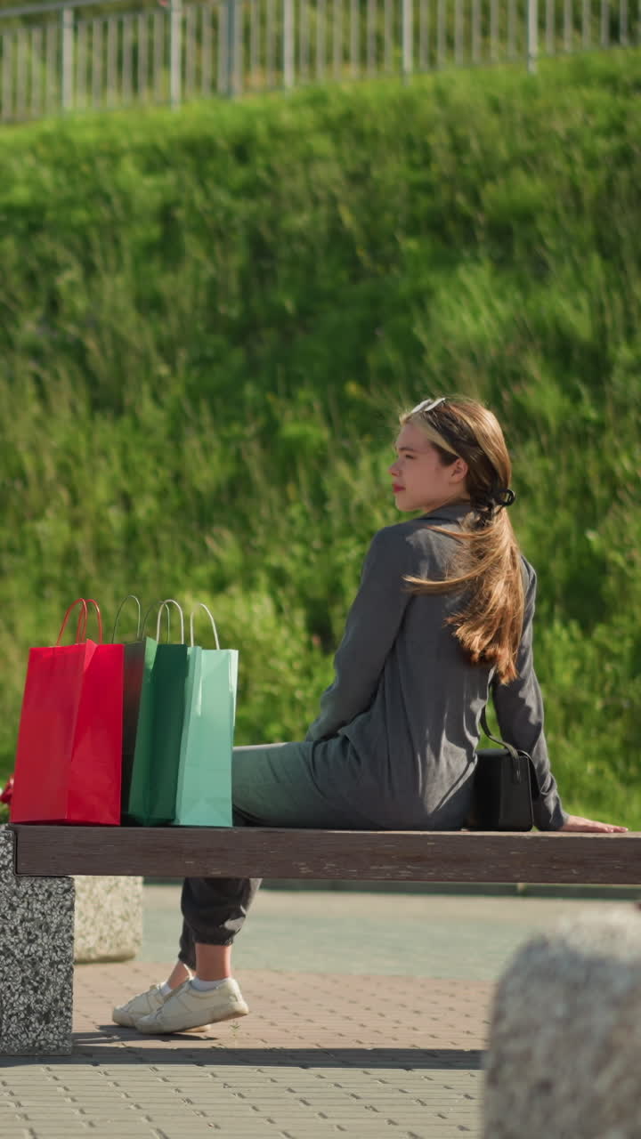 la dama coloca bolsas de compras coloridas en un banco, se sienta y se relaja bajo la luz del sol que se refleja en su cara, el fondo borroso presenta una lámpara de calle y vegetación