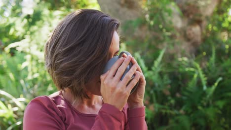 mujer caucásica sonriente bebiendo té sentada en un jardín soleado