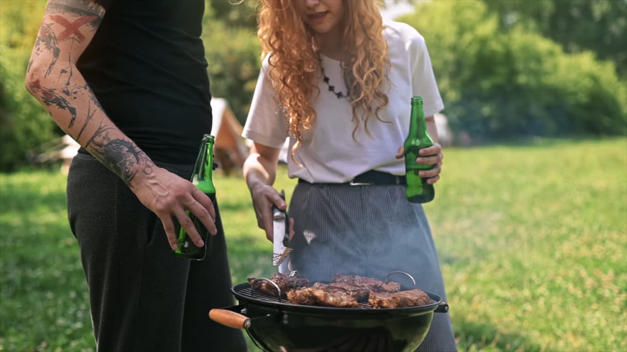 Giovane coppia che frigge carne alla griglia e beve birra. Tanto verde intorno. Glamping. Slow motion