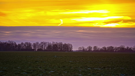 Magical-timelapse-of-yellow-sky-with-a-blanket-of-blue-clouds-beneath-and-a-green-field-with-trees-in-the-bottom-during-golden-hour