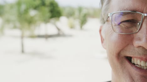 Half-portrait-of-senior-caucasian-man-in-glasses-smiling-on-beach,-in-slow-motion-with-copy-space