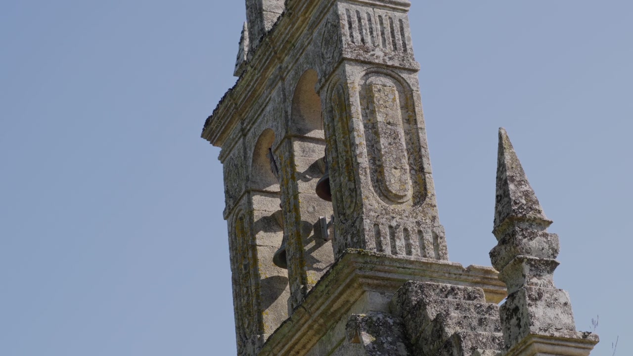 Close-up detalhado de uma torre sineira de pedra envelhecida com entalhes ornamentados sob a luz do dia brilhante, exibindo arquitetura e textura históricas.