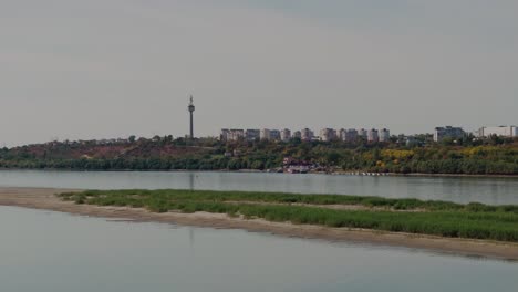 drone shot rising above the idyllic riverbank and the tranquil waters revealing a city skyline blending seamlessly with the natural landscape, sunny day 4k60fps