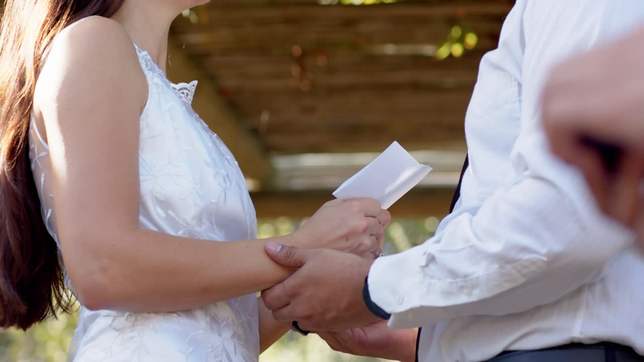 Vertical video: During wedding bride reading folded vows groom reaching clasping hands under arbor