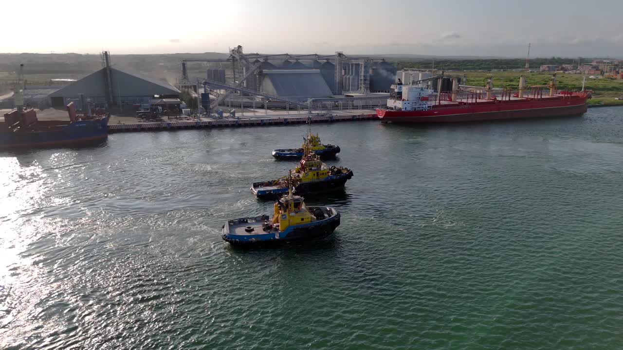 Three tugboats move through the water with the port and large vessels visible in the background.