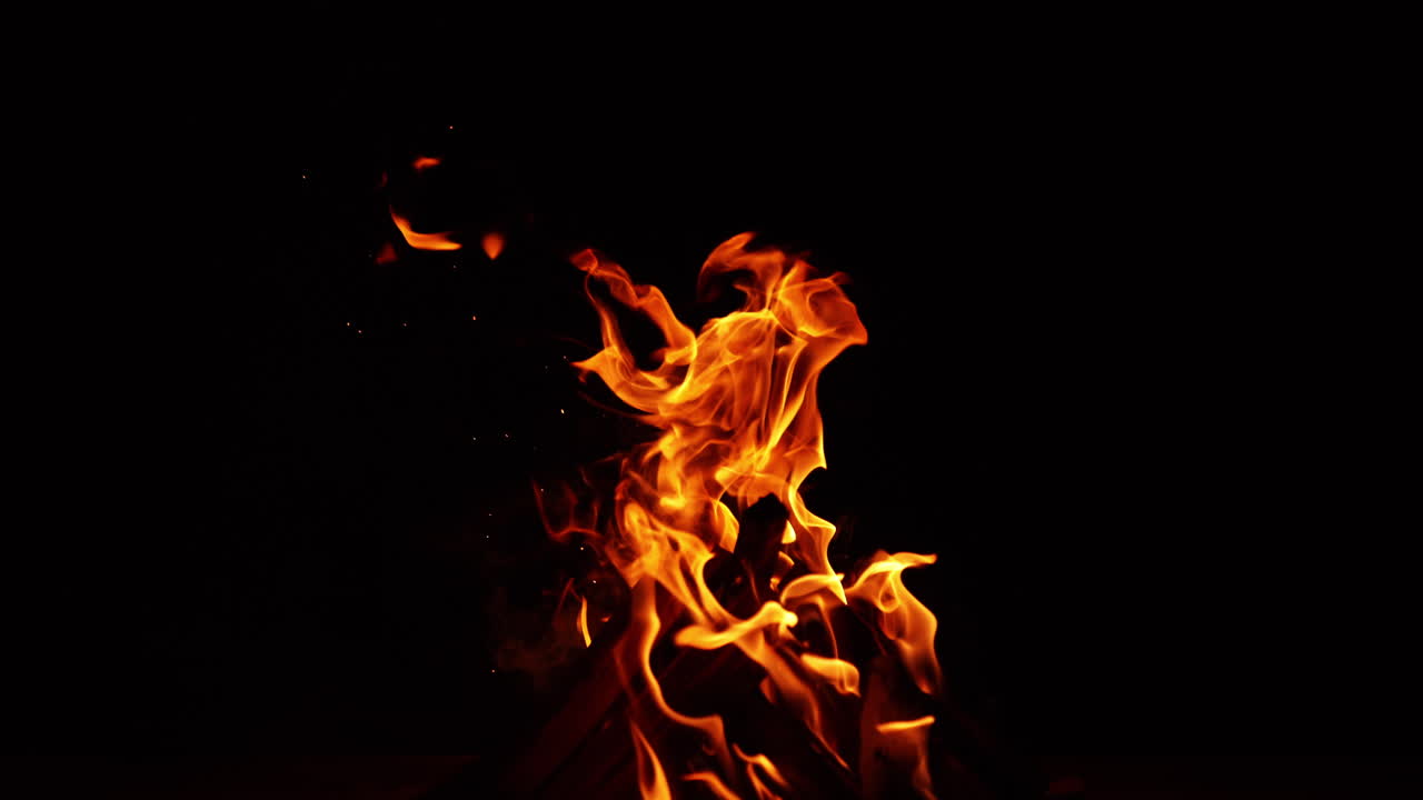 Wooden logs put in a pile burn with bright fire. Campfire blown to the left by the wind. Black backdrop.