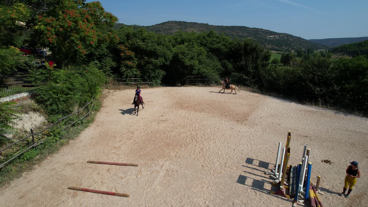 Static aerial frame shows a female rider readying for a show jumping attempt while trainer watches and another rider gallops