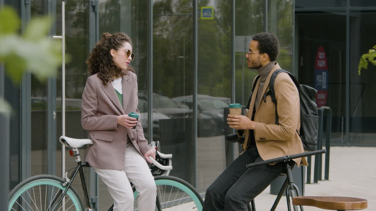 joven americano y mujer con ropa formal bebiendo café y hablando mientras se apoya en sus bicicletas en la calle 1