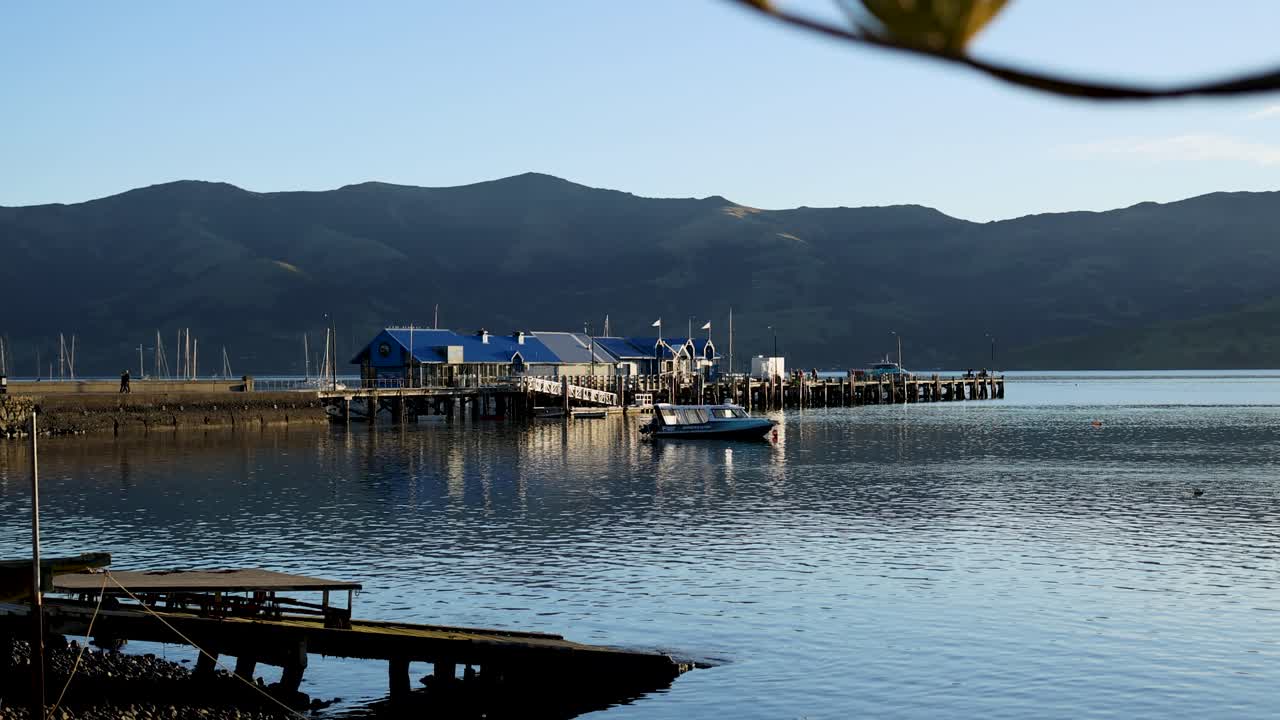 Calm waters reflect boats and mountains in Akaroa, New Zealand. Clear skies and soft lighting create a tranquil atmosphere