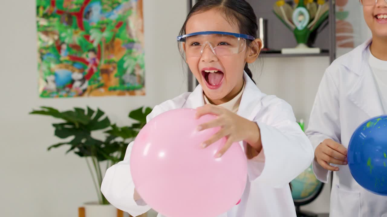Young Asian girl in safety goggles pops a pink balloon during a fun classroom experiment