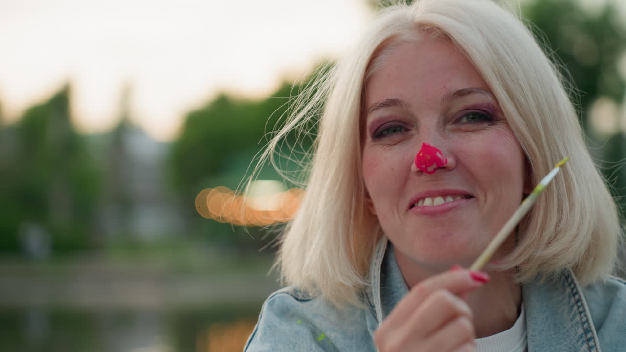 Ritratto di donna matura che tiene un pennello, con vernice brillante sul naso, che sorride calorosamente durante una giocosa sessione di pittura all'aperto vicino a un parco sul lago al tramonto, condividendo un momento artistico creativo e gioioso.