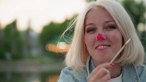 portrait of mature woman holding paint brush, with bright paint on nose, smiling warmly during playful outdoor painting session by lake park at sunset, sharing creative joyful art moment