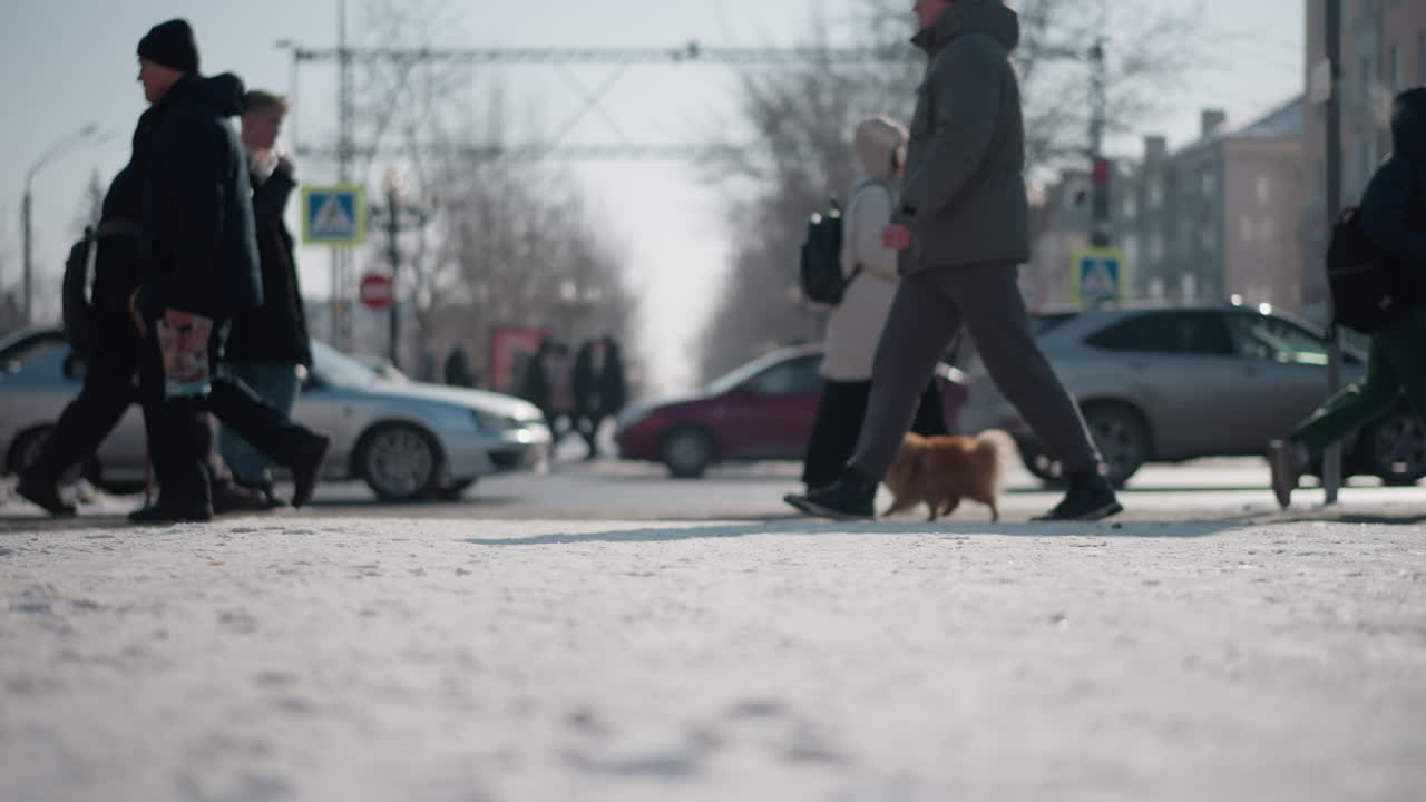 low ground view winter city scene with pedestrians in boots and coats walking while cars move in opposite lanes, snow covering street and sidewalk, urban traffic flowing through intersection